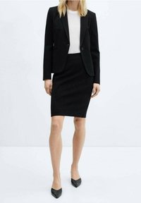 Blazer noir ajusté associé à une jupe crayon noire et un top blanc. Tissu lisse, coupe cintrée, et escarpins noirs pointus complètent le look.