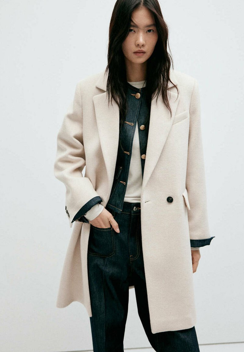 Femme aux longs cheveux noirs portant un manteau blanc par-dessus une veste en jean foncé et un pantalon, debout avec une main dans la poche sur un fond uni.