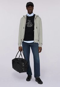 Grå huvtröja med dragkedja, svart tröja med logotyp, blå skjorta med krage, mörka jeans, svarta sneakers och en svart sportbag. En enkel lager-på-lager-outfit.