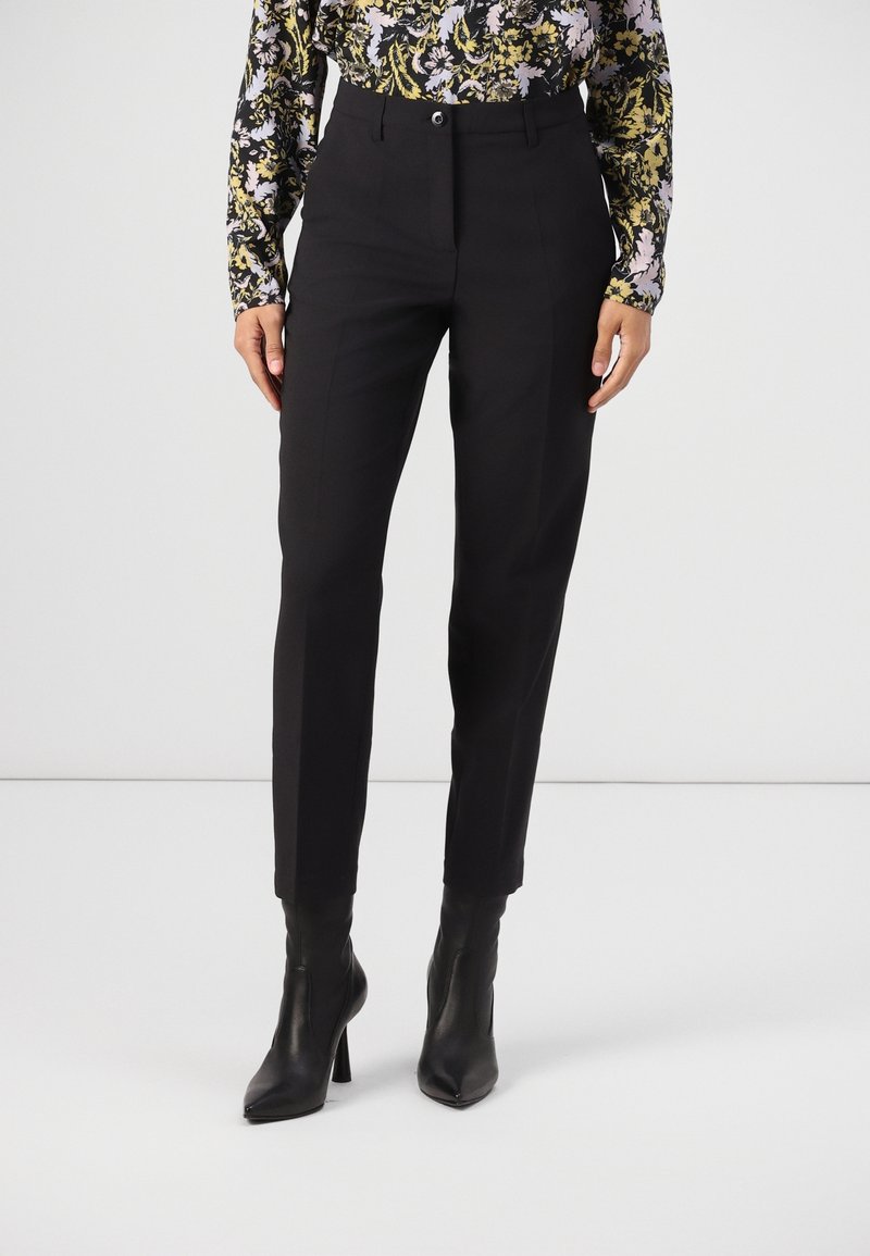 Femme portant un pantalon noir sur mesure longueur cheville, un chemisier à manches longues fleuri, et des bottines à talons noires à bout pointu, debout sur un sol blanc.