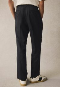Pantalon noir à rayures avec une coupe ajustée, comportant des poches avant et arrière. Associé à des baskets blanches et grises avec des accents noirs.