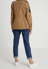 Brązowa kurtka nylonowa z regulowanym w pasie i haftowanym wzorem na lewym ramieniu. W połączeniu z ciemnoniebieskimi dżinsami z podwijanymi mankietami oraz białymi trampkami.