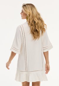 Weißes Kleid aus strukturiertem Stoff mit lockeren Ärmeln, vertikalen Nähdetails und einem gerafften Saum. Luftiges Design, ideal für warmes Wetter.