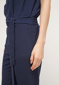 Combinaison bleu marine avec taille ceinturée, taille élastique, texture lisse et poches latérales pour une fonctionnalité supplémentaire. Gros plan sur le détail de la main.
