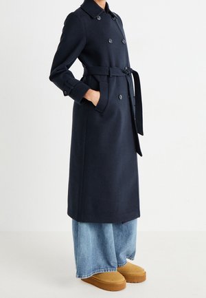 Person iført en lang, marineblå dobbeltradet frakke med bælte, lyseblå vidde-hofte jeans og beige slip-on sko, stående med hænderne i frakkens lommer.