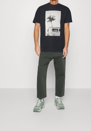 Svart t-shirt med en grafik av en palm och en VW-buss inom en vit rektangulär ram. Bärs med gröna byxor och grå sneakers.