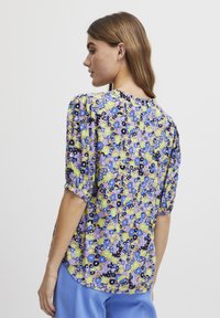 Blusa a maniche corte con effetto gonfiato, caratterizzata da un motivo floreale in blu, viola, giallo e verde su uno sfondo chiaro; scollatura rotonda; vestibilità ampia.