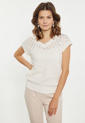 Pull en maille blanche avec un motif en losanges, col en V et manches courtes. Fini avec un ourlet côtelé, porté par-dessus un pantalon beige clair.