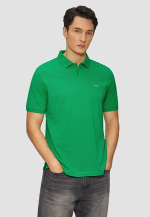Zelena majica s polo ovratnikom, izrađena od mekanog materijala, s tri gumba i malim logotipom na lijevoj strani prsiju. U kombinaciji s tamnim trapericama.
