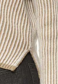 Beige-weißer gestreifter Pullover mit gerippter Textur, leicht verlängertem Rücken und seitlichen Schlitzern. Über dunklen Jeans getragen.