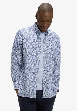 Homme portant une chemise à manches longues boutonnée avec un motif floral bleu sur un t-shirt bleu clair, regardant vers le bas avec les mains dans les poches.