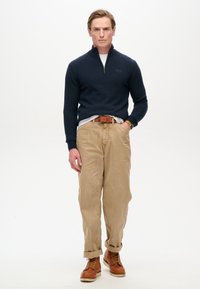 Maglione zip-up blu navale, camicia bianca, pantaloni chinos color beige, cintura in pelle marrone e stivali stringati marroni con suola crema, esposti su uno sfondo bianco.