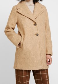 Beige wool coat with a textured surface, peak lapel, three buttons, and side pockets. Worn over brown checkered trousers.