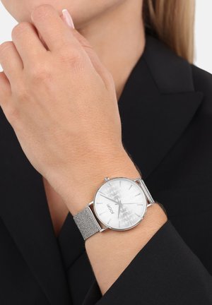 Silberne Uhr mit einem strukturierten, metallischen Mesh-Armband und einem silbernen Zifferblatt mit einem dezenten Muster, getragen am Handgelenk gegen einen dunklen Blazer.