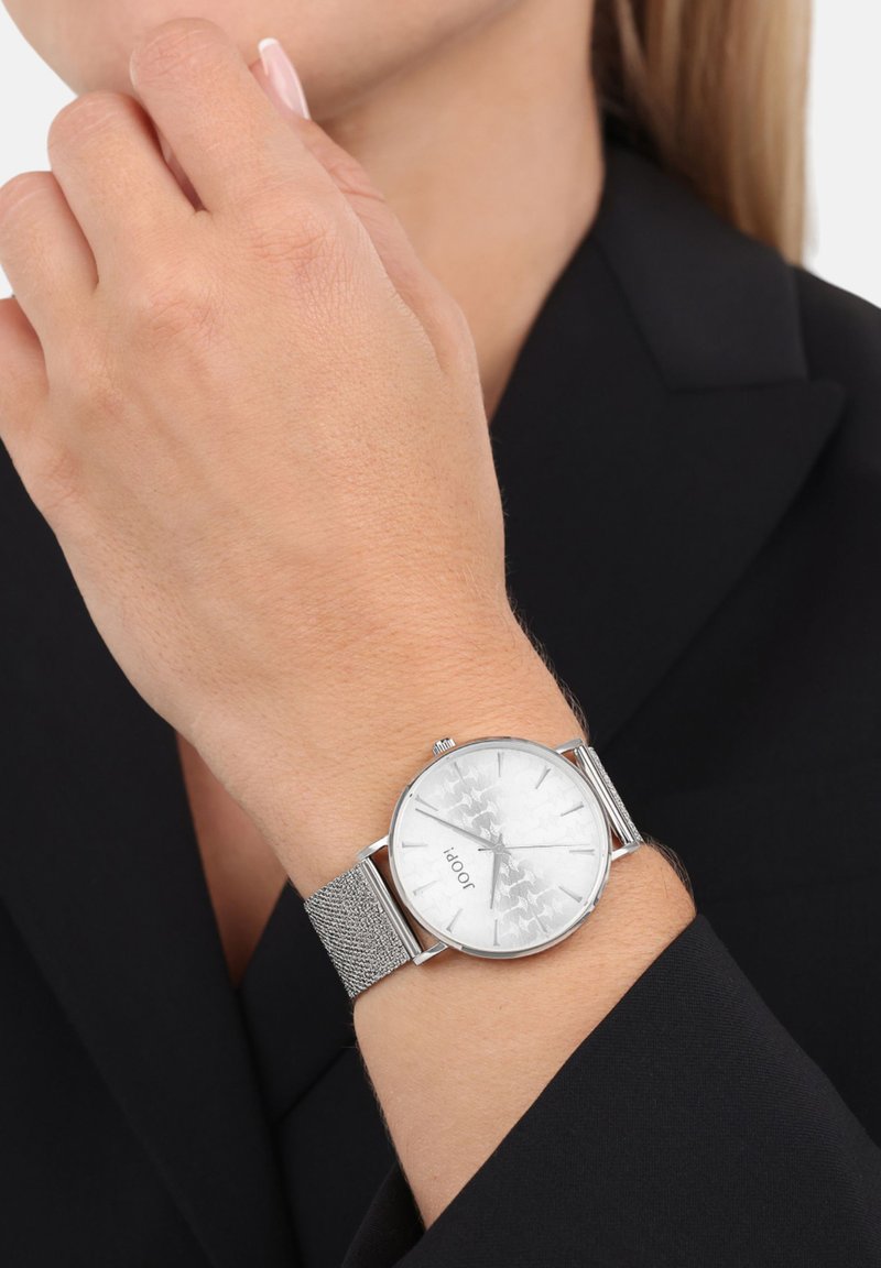 Silberne Uhr mit einem strukturierten, metallischen Mesh-Armband und einem silbernen Zifferblatt mit einem dezenten Muster, getragen am Handgelenk gegen einen dunklen Blazer.