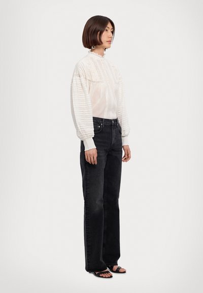 Blouse blanche texturée à manches longues avec un col haut et des détails complexes, associée à un jean noir taille haute et des sandales noires.