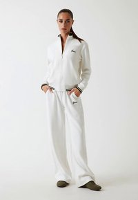 Valge soonikkoega spordidress lukuga jopaga ja elastsete varrukaotstega, millel on roheline ja must triibuline mustriga detailid ning logoga tikand mõlemal tükil.