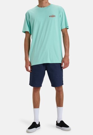 Mint green cotton t-shirt with a small logo, paired with navy shorts. White athletic socks and black slip-on shoes complete the outfit.