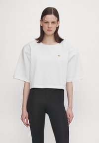 White cropped t-shirt with wide sleeves, made of cotton. Features a small gold logo on the chest, paired with black leggings.