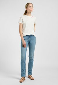 Crèmekleurig katoenen t-shirt met korte mouwen, voorzien van roze borduurwerk. Een paar lichtblauwe denim jeans en bruine sandaaltjes met bandjes.