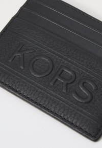 Fekete bőr kártyatartó textúrált felülettel, domború "KORS" logóval és több kártyahellyel. Minimalista dizájnnal és rendezett varrással rendelkezik.