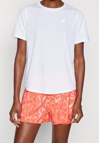 Biały sportowy t-shirt z krótkim rękawem i okrągłym dekoltem, w połączeniu z pomarańczowymi szortami sportowymi ozdobionymi falistym wzorem i drobnymi oczkami.