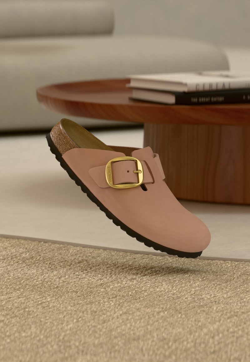 Μονό ροζ παπούτσι τύπου slip-on με χρυσή αγκράφα και μαύρη σόλα να αιωρείται πάνω από υφασμένο μπεζ χαλί, με ξύλινο τραπεζάκι καφέ και βιβλία στο φόντο.