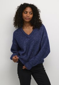 Pulover tricotat cu decolteu în V, de culoare albastru închis, cu o croială lejeră, material texturat și umeri căzuți, împreună cu pantaloni negri.