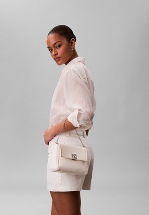 Femme aux cheveux foncés attachés, portant une blouse blanche et un short, tenant un sac à main blanc texturé avec une chaîne et une fermeture argentées.