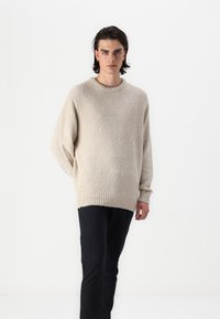Beige textured sweater with a round neckline and ribbed hem, paired with black pants. The sweater features loose, oversized sleeves.