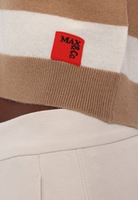 Gros plan sur un tricot rayé beige et blanc avec une étiquette carrée rouge cousue sur le bord, associé à un pantalon en tissu clair.