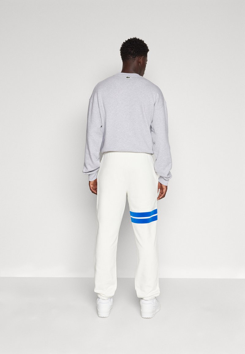 Felpa grigia e pantaloni della tuta bianchi con strisce orizzontali blu sulla gamba sinistra. I pantaloni hanno polsini elasticizzati e una vestibilità comoda.