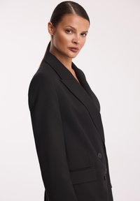 Czarna marynarka o dopasowanym kroju, z ostrymi klapami, guzikami z przodu i bocznymi kieszeniami, wykonana z gładkiego materiału o matowym wykończeniu.