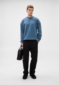 Ανοιχτό μπλε φούτερ με στρογγυλή λαιμόκοψη, συνδυασμένο με μαύρο στενό παντελόνι. Το μοντέλο κρατά μια μαύρη δομημένη τσάντα. Απλός, μοντέρνος σχεδιασμός.