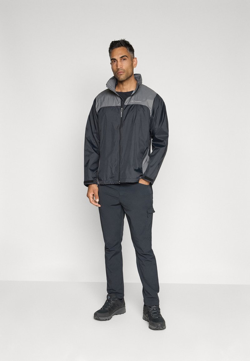 Chaqueta negra resistente al agua con detalles grises, cierre de cremallera, cuello alto y puños elásticos, combinada con pantalones cargo negros a juego y botas.