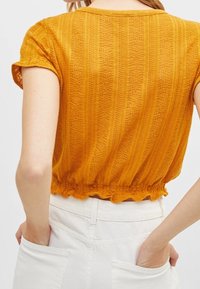 Kvinna iklädd en senapsgul texturerad kortärmad topp med volangkant, instoppad i högmidjade vita byxor.