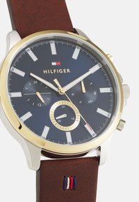 Orologio da uomo con quadrante blu navy, cassa in acciaio inossidabile color oro, cinturino in pelle marrone e tre sottocronografi. Presenta marker delle ore bianchi e logo.