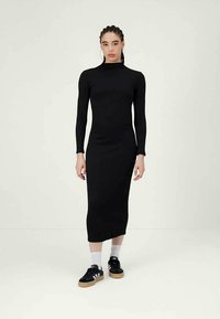 Γυναίκα με πλεξούδες που φοράει ένα μαύρο μίντι φόρεμα με μακριά μανίκια, άσπρες κάλτσες και μαύρα αθλητικά παπούτσια Adidas, στέκεται σε λευκό δάπεδο ενάντια σε ένα απλό φόντο.