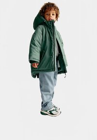 Zelená prošívaná bunda s kapucí, pod ní světle šedé tričko, modré džíny a zeleno-bílé tenisky. Bunda má zapínání na patentky.