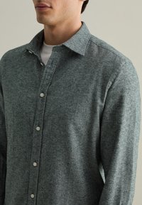 Camicia grigia a spina di pesce con una superficie morbida, colletto e bottoni visibili lungo la patta frontale.