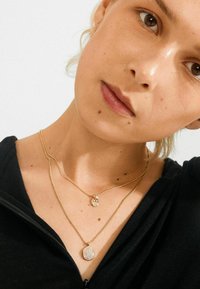 Close-up van een persoon die twee lagen gouden halskettingen met ronde kristallen hangers draagt boven een zwart met rits top.