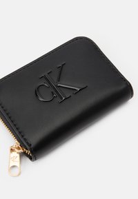 Black leather wallet featuring a glossy embossed "CK" logo, gold zipper hardware, and smooth texture with stitched edges.