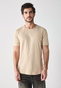 Beige lyhythermainen t-paita pyöreällä kaula-aukolla, valmistettu kevyestä kankaasta. Sisältää hienovaraisen tekstuurin, yhdistettynä tummiin farkkuihin.