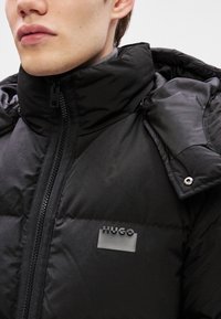 Black padded jacket featuring a high collar, zip closure, and logo detail. Material appears glossy with a smooth texture, accentuated by a detachable hood.