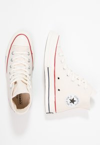 Béžové vysoké tenisky s bílými gumovými podrážkami, červenými a modrými akcentními pruhy a kovovými oky. Obsahují klasické logo Converse All-Star.