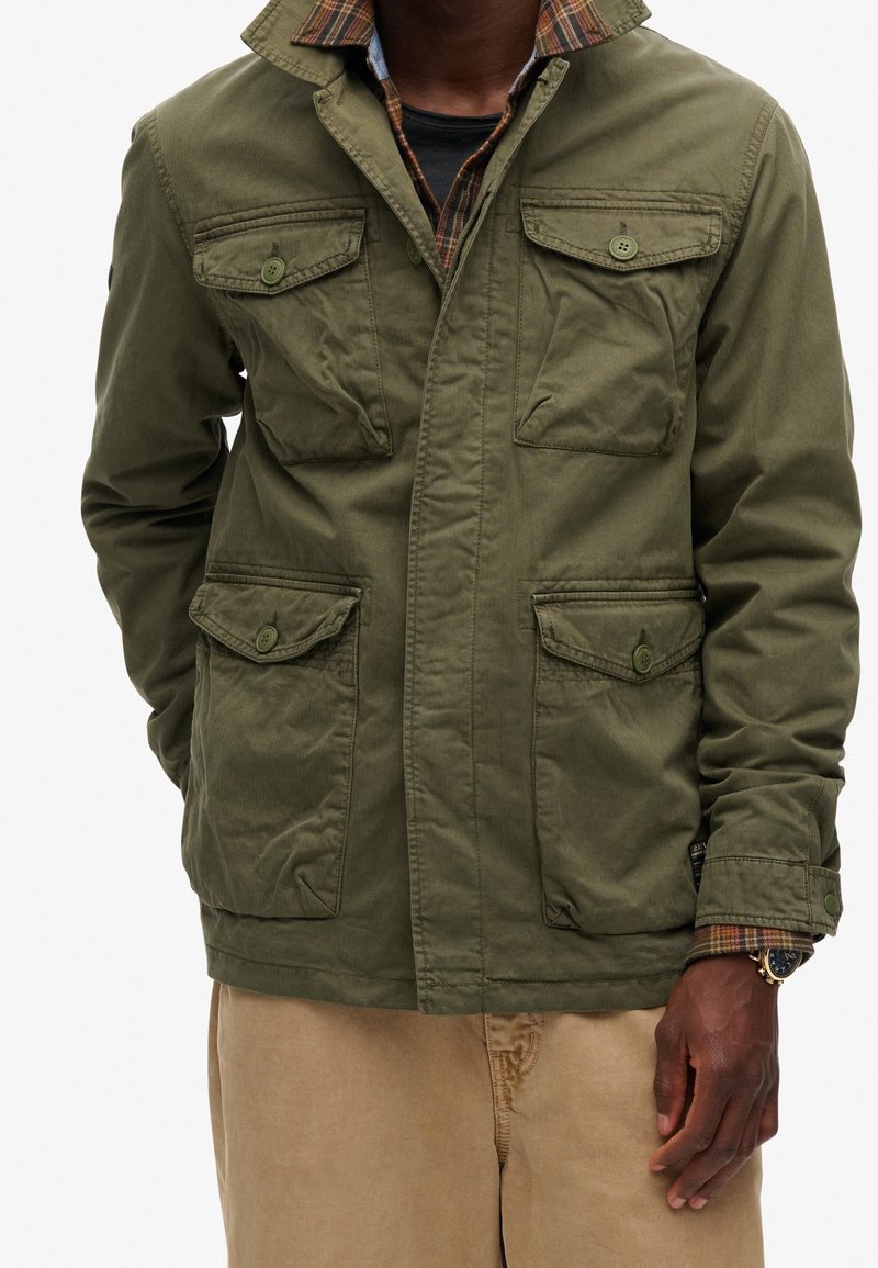 Personne à la peau foncée portant une veste boutonnée vert olive sur une chemise à carreaux et un pantalon beige, avec une montre au poignet gauche.