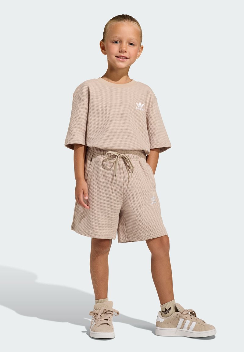 Bež rebrasti pamučni komplet majica kratkih rukava i kratkih hlača s vezicama u struku. Bijeli logotipi na oba dijela. Uparen s bež tenisicama i čarapama.