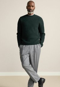 Dunkelgrüner gerippter Pullover mit rundem Halsausschnitt, kombiniert mit hellgrauen, gefalteten Hosen und schwarzen Loafers, vor einem neutralen Hintergrund.