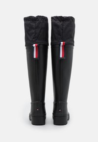 Bottes en caoutchouc noires montant jusqu'au genou avec un dessus en tissu noir matelassé. Présente des rayures accentuées rouges, blanches et bleues à l'arrière. Semelle texturée.