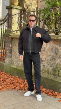 Crna bomber jakna s podstavom i prednjim zatvaračem, ispod siva majica, crne traperice i bijele tenisice. U pozadini je kameni zid i biljke.